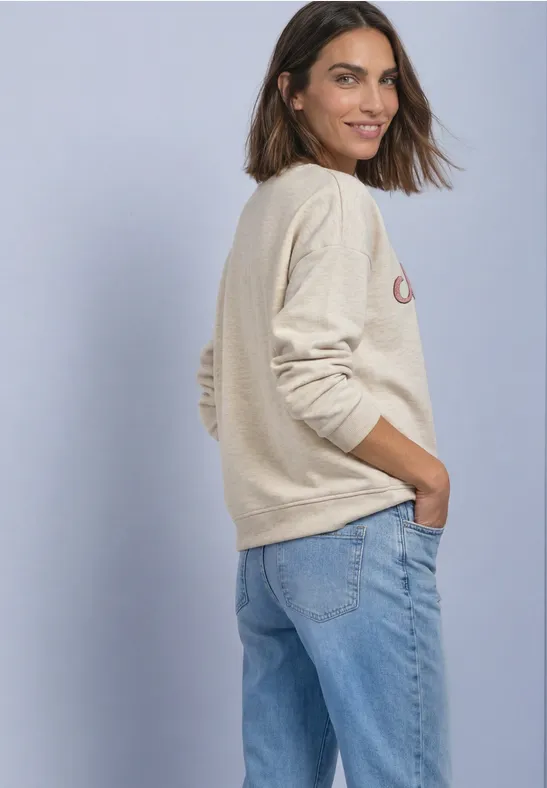 Femme souriante de profil en pull beige et jean bleu clair, les mains dans les poches.