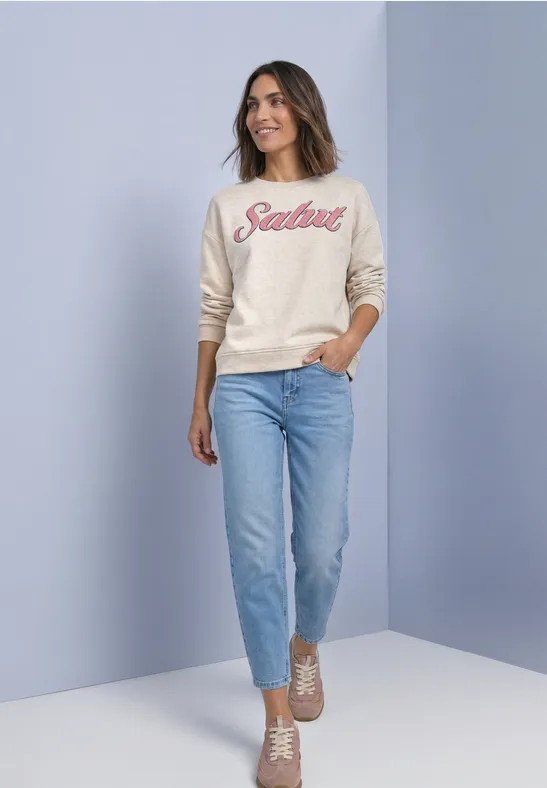 Une femme en jean, sweat clair 'Salut' et baskets beiges devant un mur pastel.