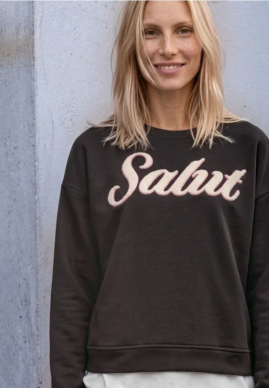 Jeune femme blonde souriante appuyée contre un mur, portant un pull noir orné de l'inscription ‘Salut’.