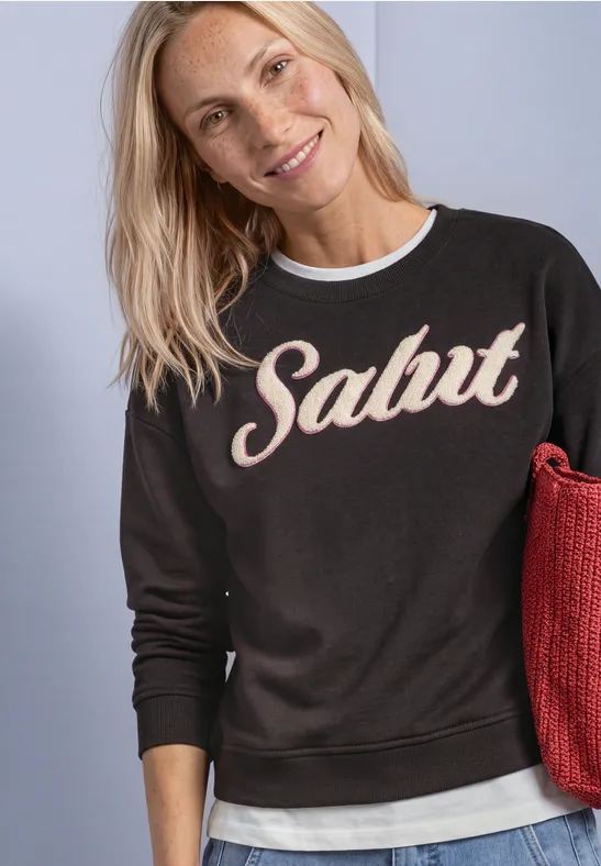 Une femme blonde souriante porte un sweat-shirt noir avec l’inscription « Salut » et tient un sac rouge.