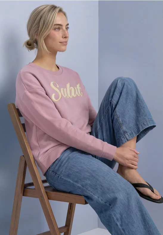Jeune femme assise de côté sur une chaise pliante, sweat rose “Salut”, jean large et tongs sur fond pastel.