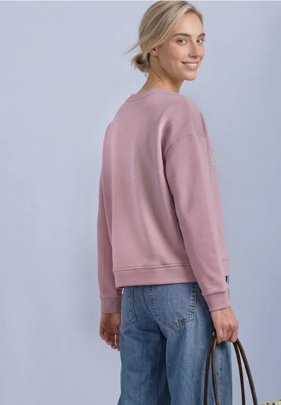 Une femme en pull rose et jean bleu regarde par-dessus son épaule en tenant un sac à main.