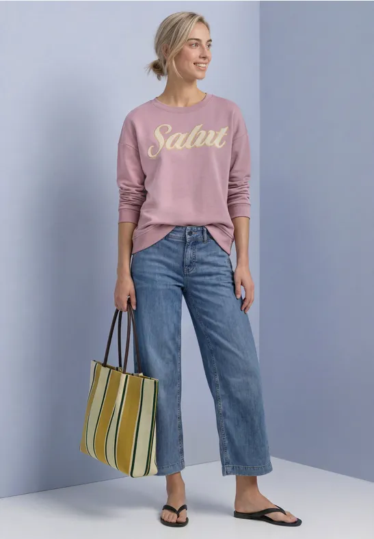 Femme en pull rose "Salut", jean ample, sac rayé et sandales.