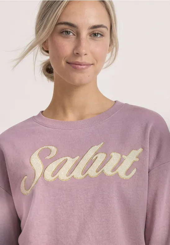 Jeune femme portant un sweat rose avec l'inscription dorée 'Salut', sourire discret.