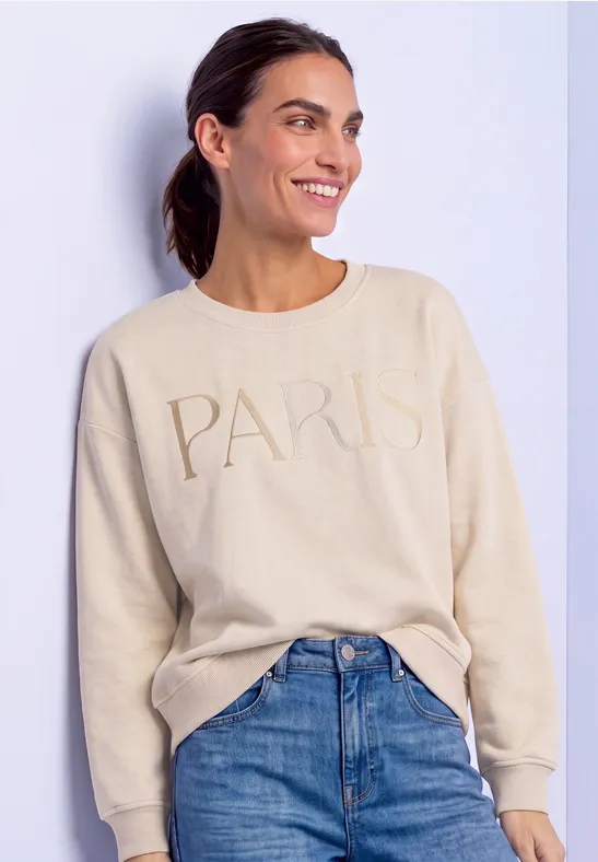 Femme souriante en pull beige 'PARIS' et jean, regard détourné.