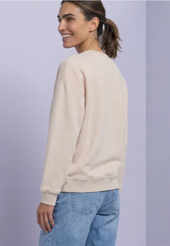 Jeune femme en pull beige et jean, vue latérale, souriante devant un fond violet.