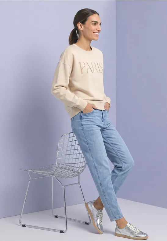 Femme en sweat beige, jean clair et baskets argentées appuyée sur une chaise en fil métallique devant un fond pastel.