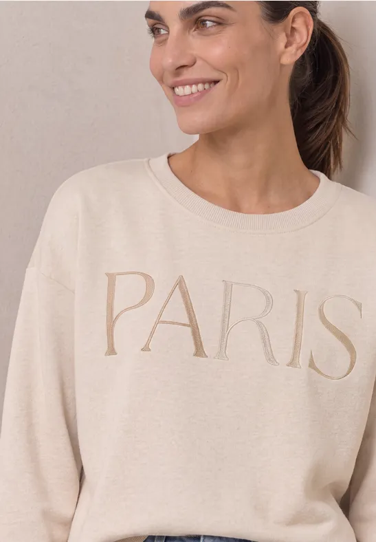 Une femme souriante portant un pull beige avec l’inscription PARIS.