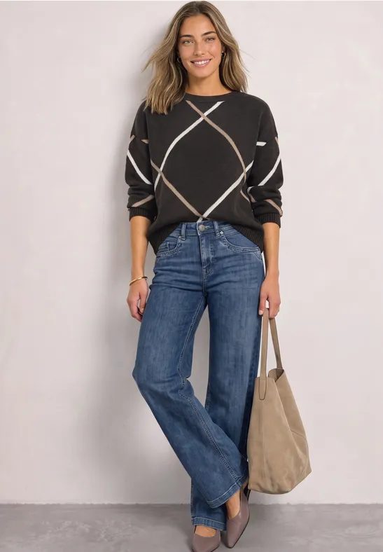Vrouw in zwarte trui met ruitpatroon, blauwe jeans en beige tas, glimlachend voor een lichte muur.