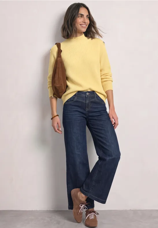 Vrouw leunt lachend tegen muur, draagt gele trui, wijde jeans, bruine schoenen en tas.