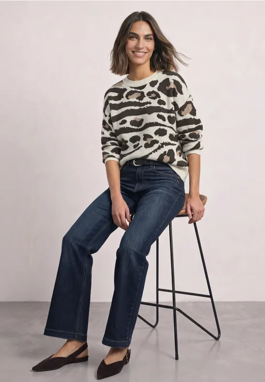 Vrouw zit op kruk in animalprint trui, rechte donkerblauwe jeans en zwarte pumps, ze lacht.