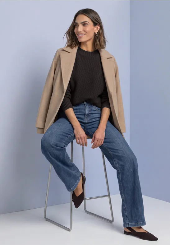 Vrouw zit ontspannen op kruk, draagt beige blazer over zwart topje, wijdvallende jeans en puntige schoenen.