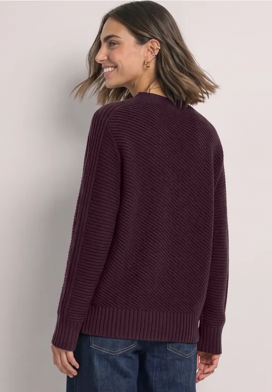 Eine Frau mit schulterlangen Haaren trägt einen weinroten Strickpullover und lächelt seitlich.