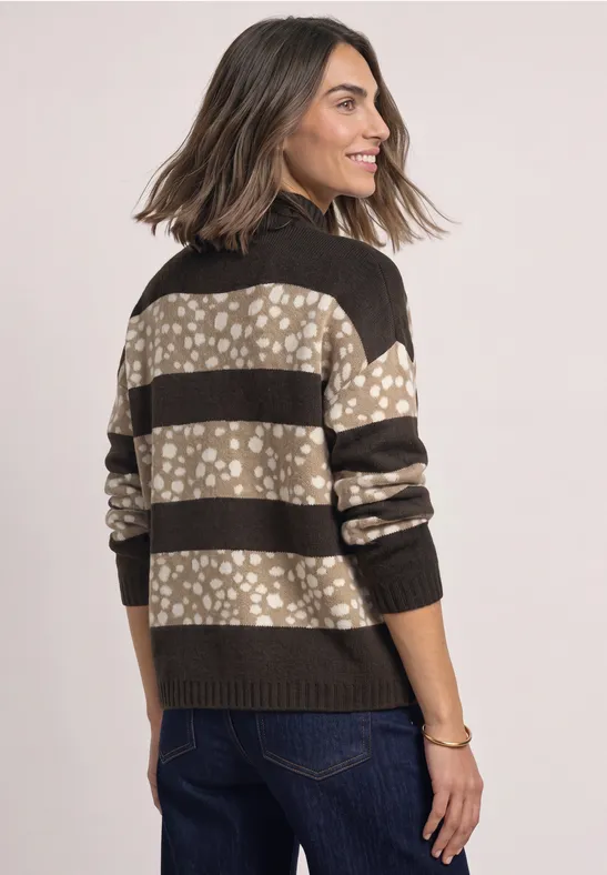 Vrouw in gestreepte beige-zwart trui met bloemmotieven en jeans, tegen lichte achtergrond.