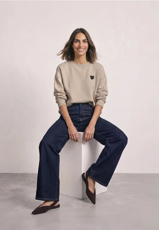 Femme souriante portant un sweat beige et un jean évasé, assise sur un tabouret blanc devant un fond clair.
