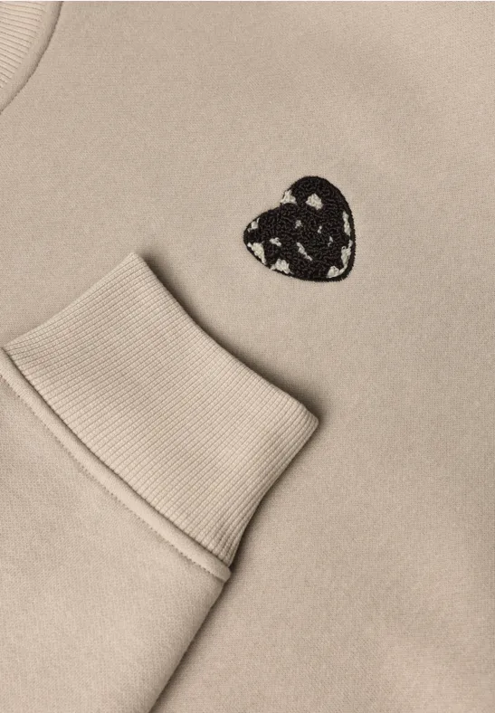 Sweat beige avec poche et petit écusson cœur noir orné de motifs triangulaires blancs.