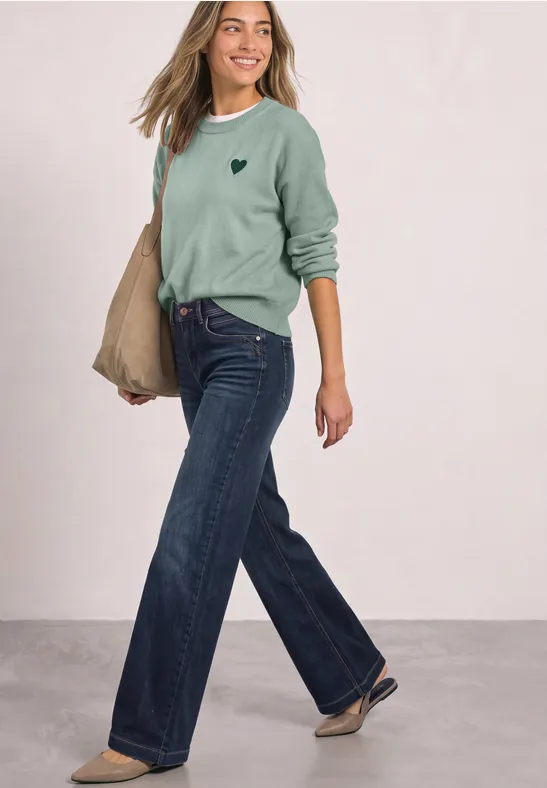 Une femme souriante porte un pull vert, un jean large et un sac beige.