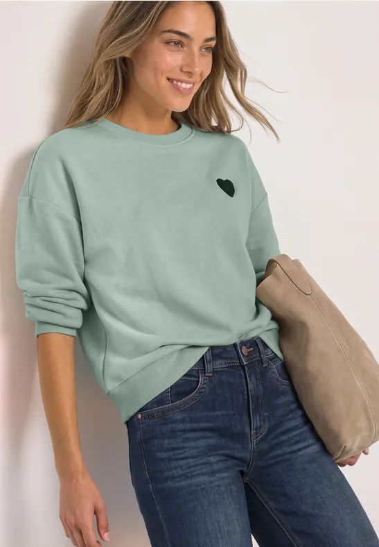 Femme en sweat vert à motif cœur et jeans, tenant un manteau beige, souriante devant un mur blanc.