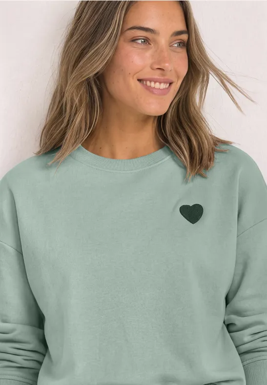 Femme souriante en sweat vert clair avec un petit cœur noir sur la poitrine.