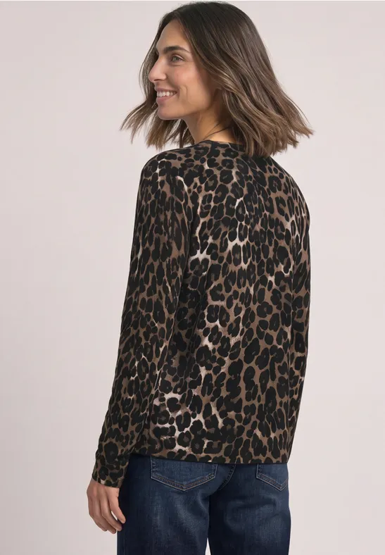 Een vrouw draagt een bruine luipaardtrui en een blauwe jeans, kijkt over haar schouder en lacht.