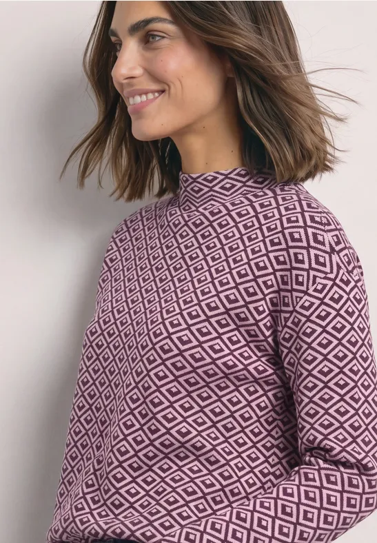 Eine Frau in einem lila Pullover mit geometrischem Muster, lächelnd, seitlich stehend.
