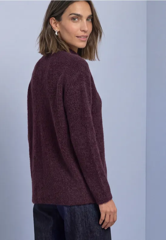 Frau in dunkelrotem Strickpullover und Jeans vor neutralem Hintergrund, blickt über die Schulter.