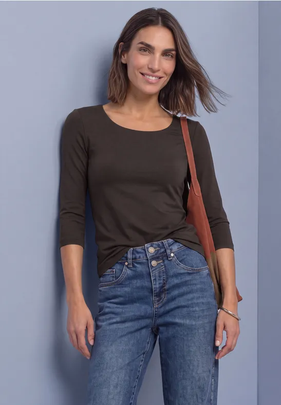 Brünette Frau in schwarzem 3/4-ärmligem Shirt und blauen Jeans lehnt lächelnd an einer Wand.