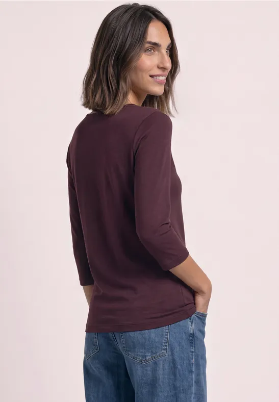 Frau in weinrotem 3/4-Arm-Shirt und Jeans, lächelt über die Schulter, Hände in den Taschen.