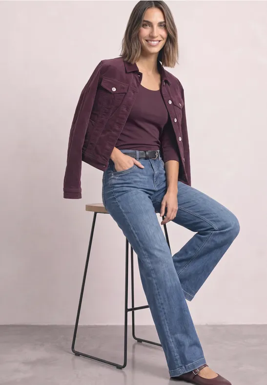 Eine Frau lehnt sich lässig auf einen Barhocker, trägt violetten Blazer, lila Top und blaue Jeans.