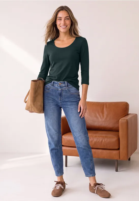 Junge Frau steht in einem hellen Raum, trägt grüne Bluse, blaue Jeans, braune Schuhe und hält eine Tasche.