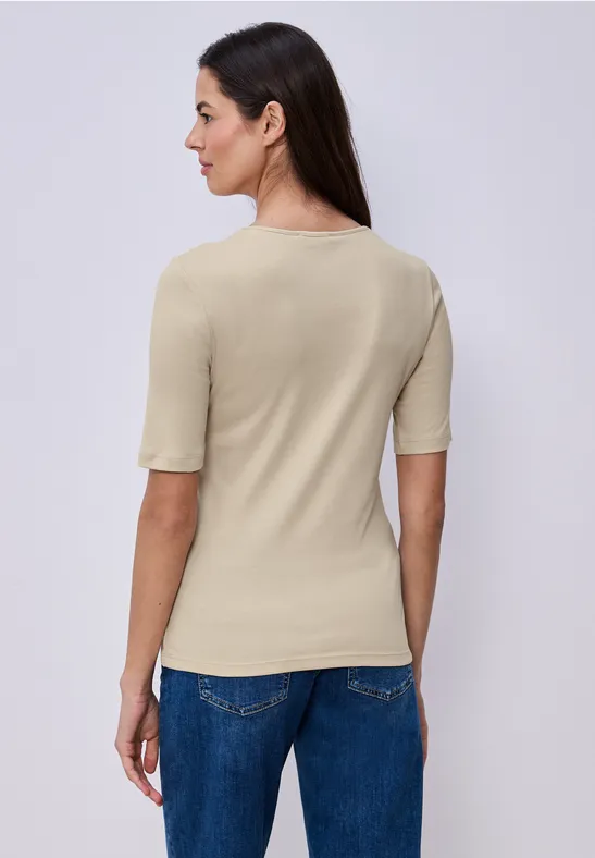 Vrouw met beige top met korte mouwen en blauwe jeans, zijwaarts zichtbaar tegen witte achtergrond.