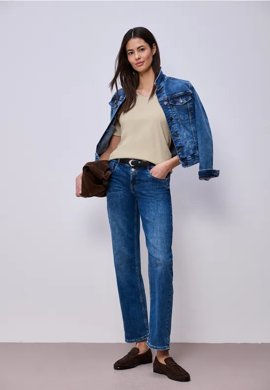 Vrouw in denim: spijkerjasje en jeans, beige top en bruine loafers, met tas voor witte achtergrond.