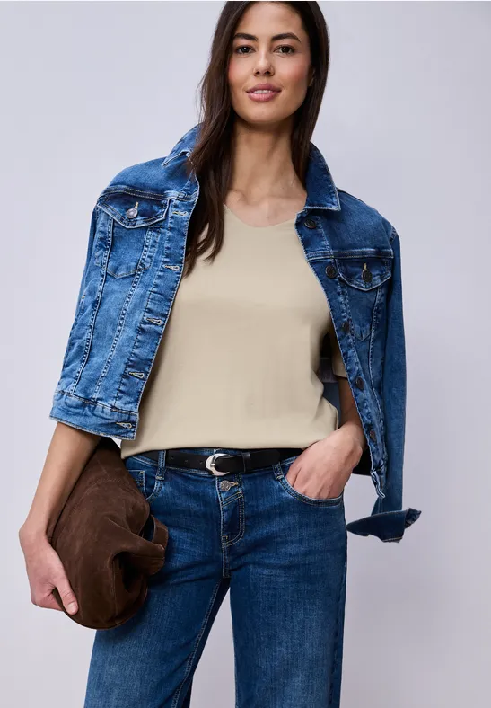 Vrouw in beige top, blauwe jeans, spijkerjack op schouders en een bruine clutch.