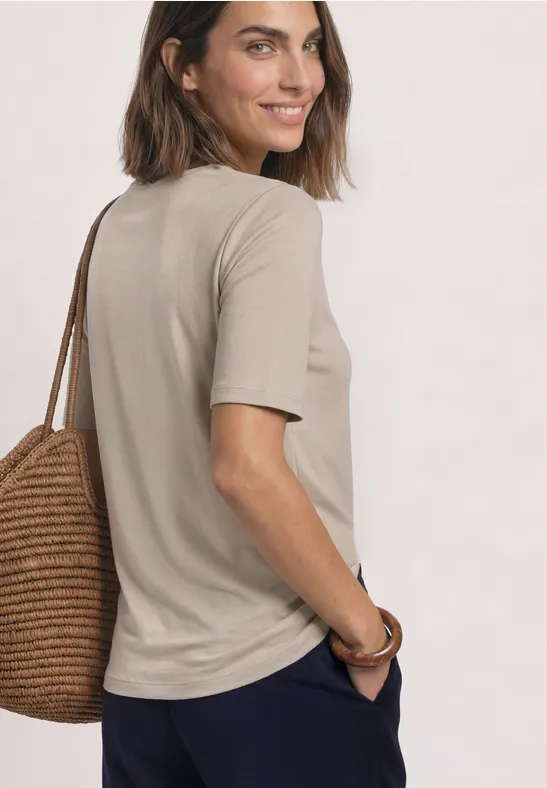 Femme en T-shirt beige avec sac en paille, souriante, regard par-dessus l'épaule sur fond clair.