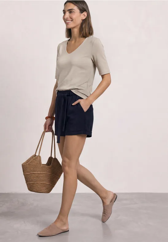 Femme en t-shirt beige et short noir, tenant un sac en osier, chaussures plates, sur fond clair.