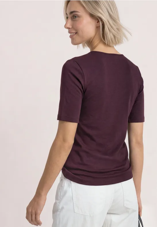 Rückansicht einer Frau in einem kurzärmligen, dunkelroten Shirt und hellen Hosen vor hellem Hintergrund.