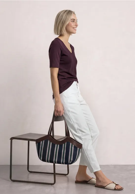 Eine Frau in weißer Hose und dunklem Oberteil hält eine gestreifte Tasche und blickt seitlich.