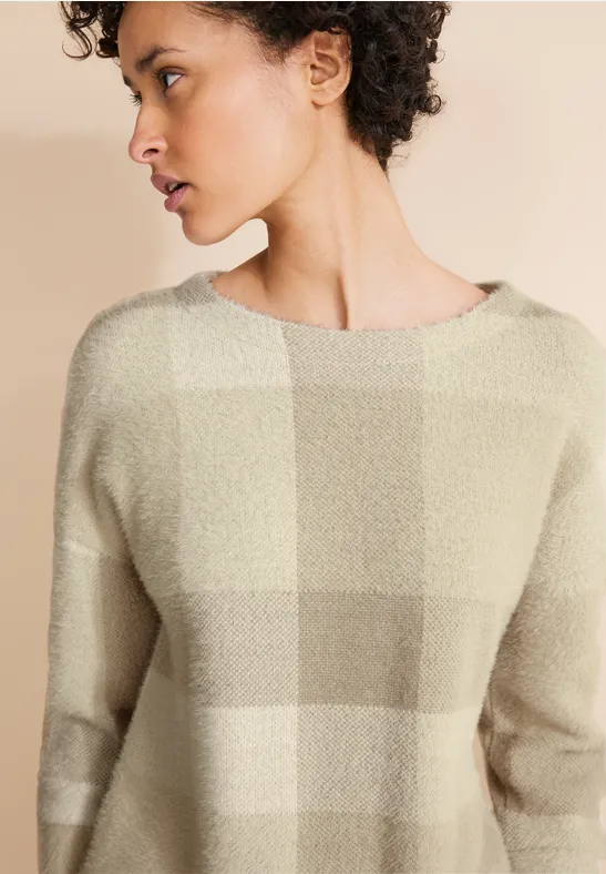 Vrouw met krullend haar draagt beige geruite trui en kijkt opzij.