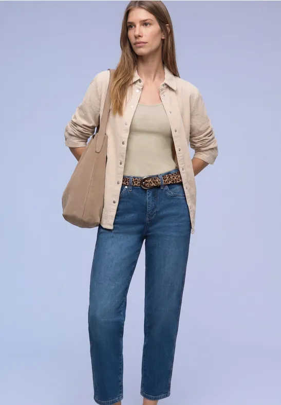 Frau in lässiger Jeans, beigem Hemd über einem olivgrünen Top, mit Gürtel und passender Tasche.