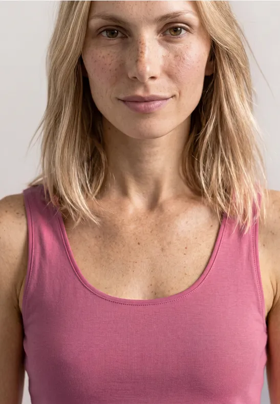 Vrouw met halflang blond haar draagt een mouwloos roze shirt voor een lichte achtergrond.