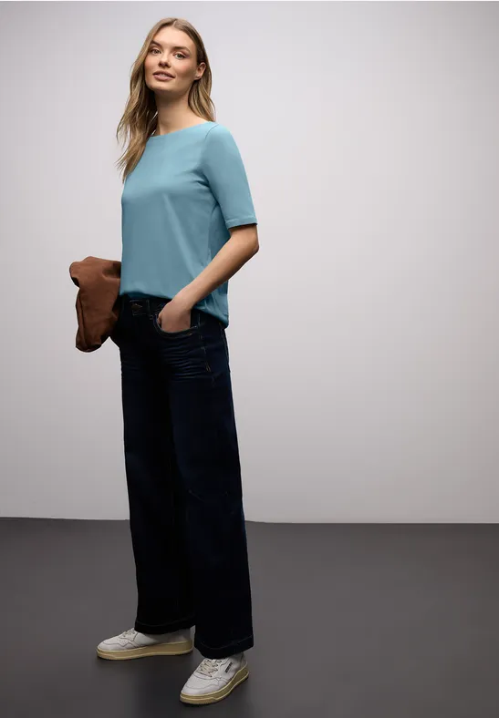 Femme en haut bleu clair, jean large foncé, baskets blanches et porte un sac marron.