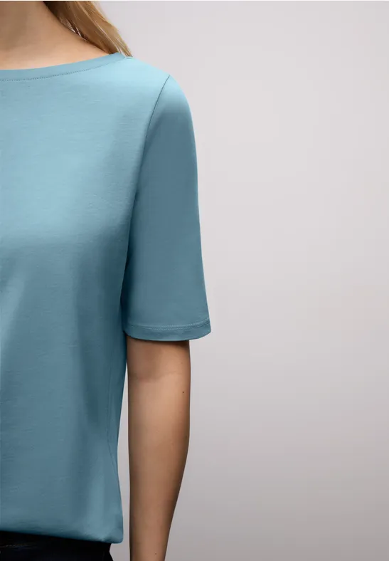 Une personne porte un t-shirt turquoise à manches courtes devant un fond neutre clair.