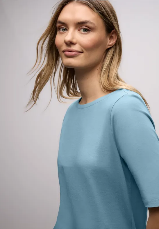 Une femme vue de côté porte un haut bleu clair à manches mi-longues en tissu doux, sur fond neutre.