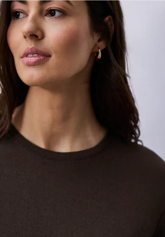 Femme aux cheveux bruns longs portant un haut marron foncé et une petite boucle d'oreille dorée