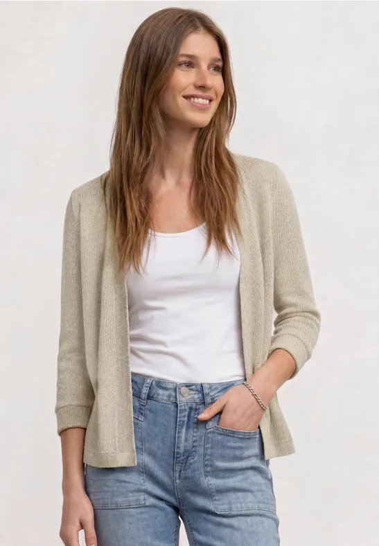 Vrouw in beige vest, witte top en blauwe jeans, staand voor een lichte achtergrond met één hand in haar zak.