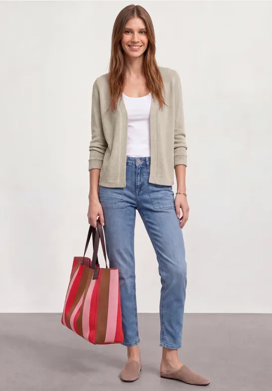 Vrouw in wit hemd, beige vest, blauwe jeans en platte schoenen, met een rood gestreepte tas.