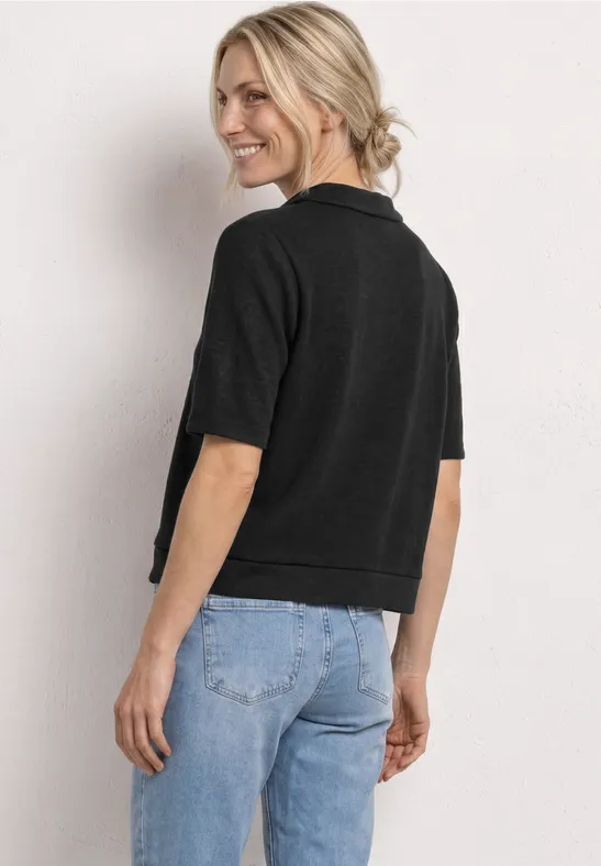 Vrouw van achter, draagt zwarte kortemouwtrui en lichtblauwe jeans, tegen neutrale achtergrond.