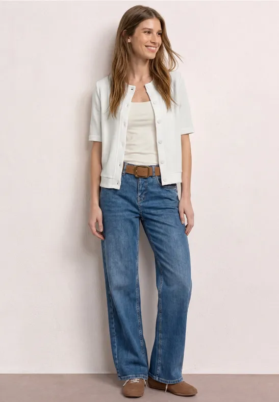 Vrouw met wit kort jasje, wit topje, blauwe jeans, bruine riem en loafers.