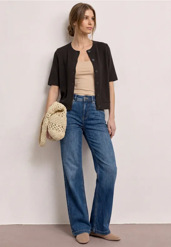 Vrouw in beige top, zwart kort jasje, wijde blauwe jeans en met een gevlochten tas.