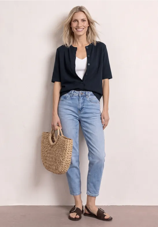 Vrouw in marineblauw vest, witte top, lichtblauwe jeans, zwarte sandalen en strooien tas.